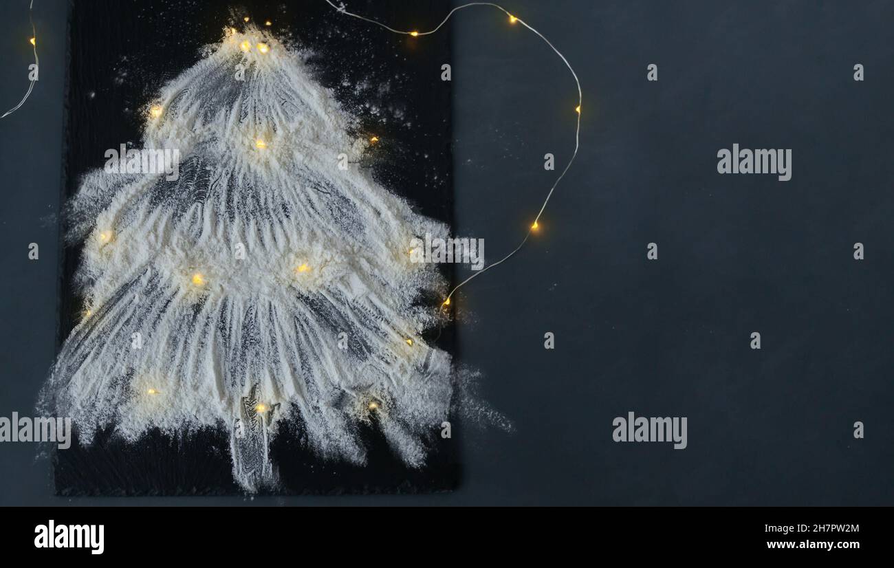 Weihnachtsbaum mit Mehlschnee geschmückt mit Weihnachtsbeleuchtung auf einem schwarzen Schiefer Food Board Hintergrund. Draufsicht. Speicherplatz kopieren. Horizontale Ausrichtung. Stockfoto