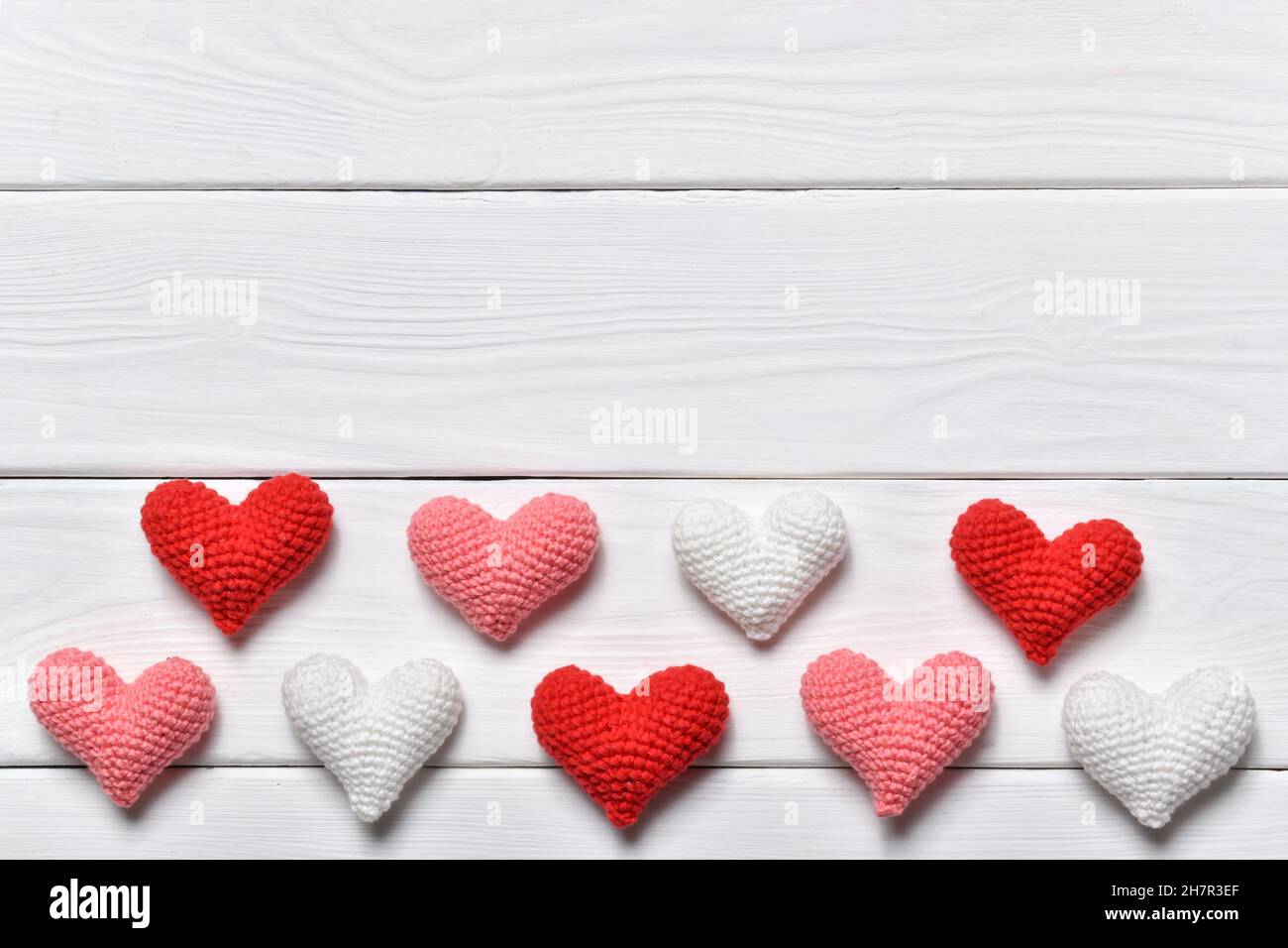 Der Hintergrund des Valentinstag. Kleine gestrickte Herzen auf weißem Holzhintergrund. Herzen in drei Farben. Minimalismus ist Liebe. Stockfoto