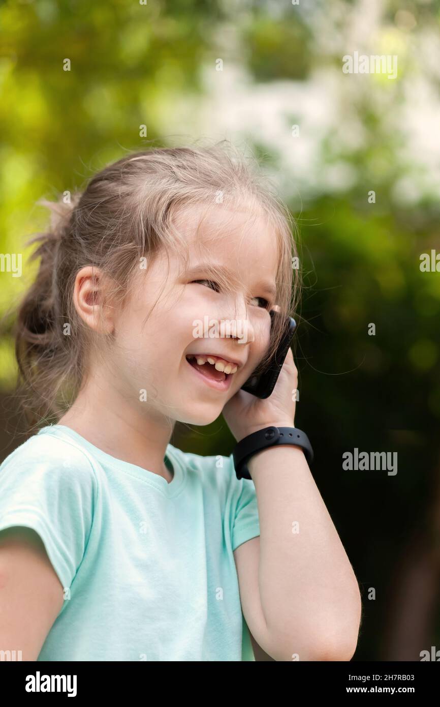 Fröhliches, fröhliches Kind im Grundschulalter, ein Mädchen, das auf ihrem Handy spricht, ein Smartphone mit Smartwatch neben dem Ohr hält und lacht. Im Freien Stockfoto