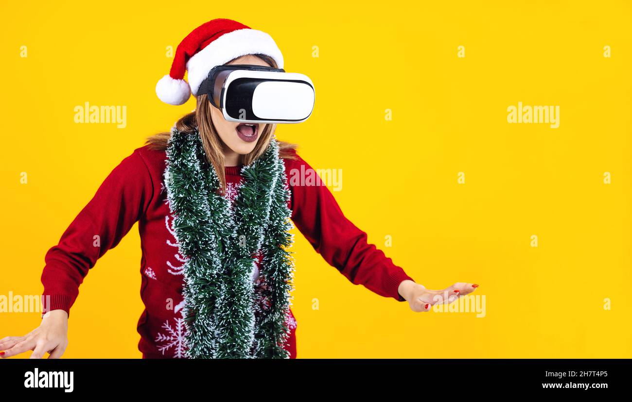 Junge lateinische Frau Gamer in Virtual Reality-Headset-Gadget trägt Weihnachtsmütze und Weihnachts-Pullover auf gelbem Hintergrund. Weihnachten und Winter vr Techno Stockfoto