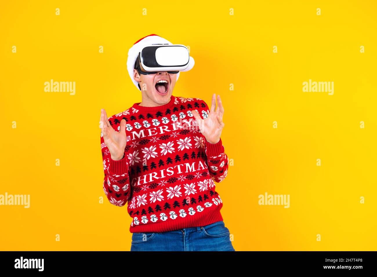 Junger latein Mann Gamer in Virtual Reality Headset Gadget trägt Weihnachtsmütze und Weihnachtspullover auf gelbem Hintergrund. Weihnachten und Winter vr technolo Stockfoto