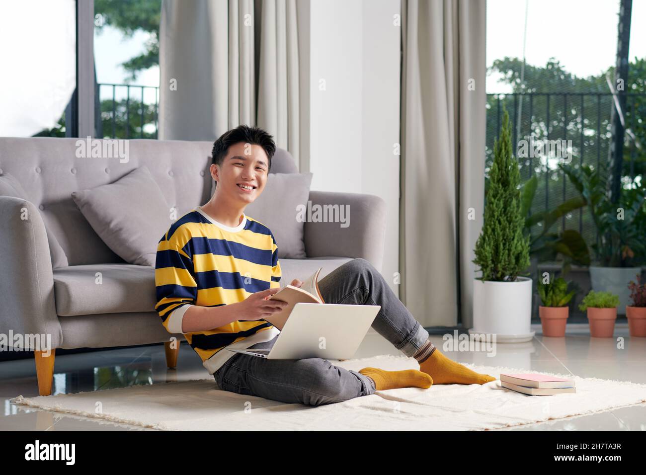 Freiberuflicher Arbeitstag. Lächelnder, tausendjähriger Student, der von zu Hause aus auf dem Teppich im Wohnzimmer sitzt und studiert. Stockfoto