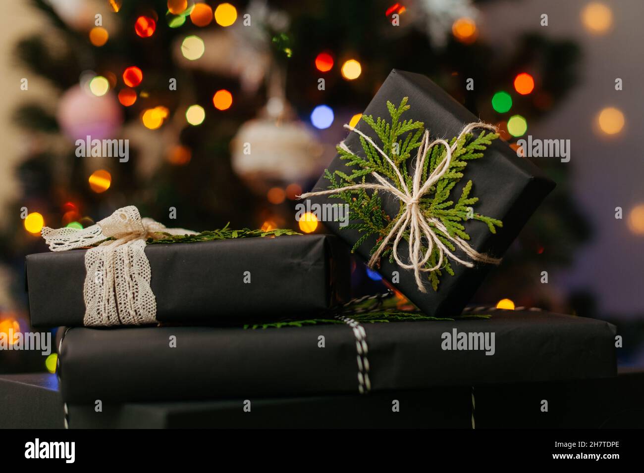 Schöne gemütliche Geschenkboxen in schwarzem Papier gewickelt, die auf grünem Textil liegen, defokussiertes Licht auf Weihnachtsbaum im Hintergrund. Nahaufnahme Stockfoto