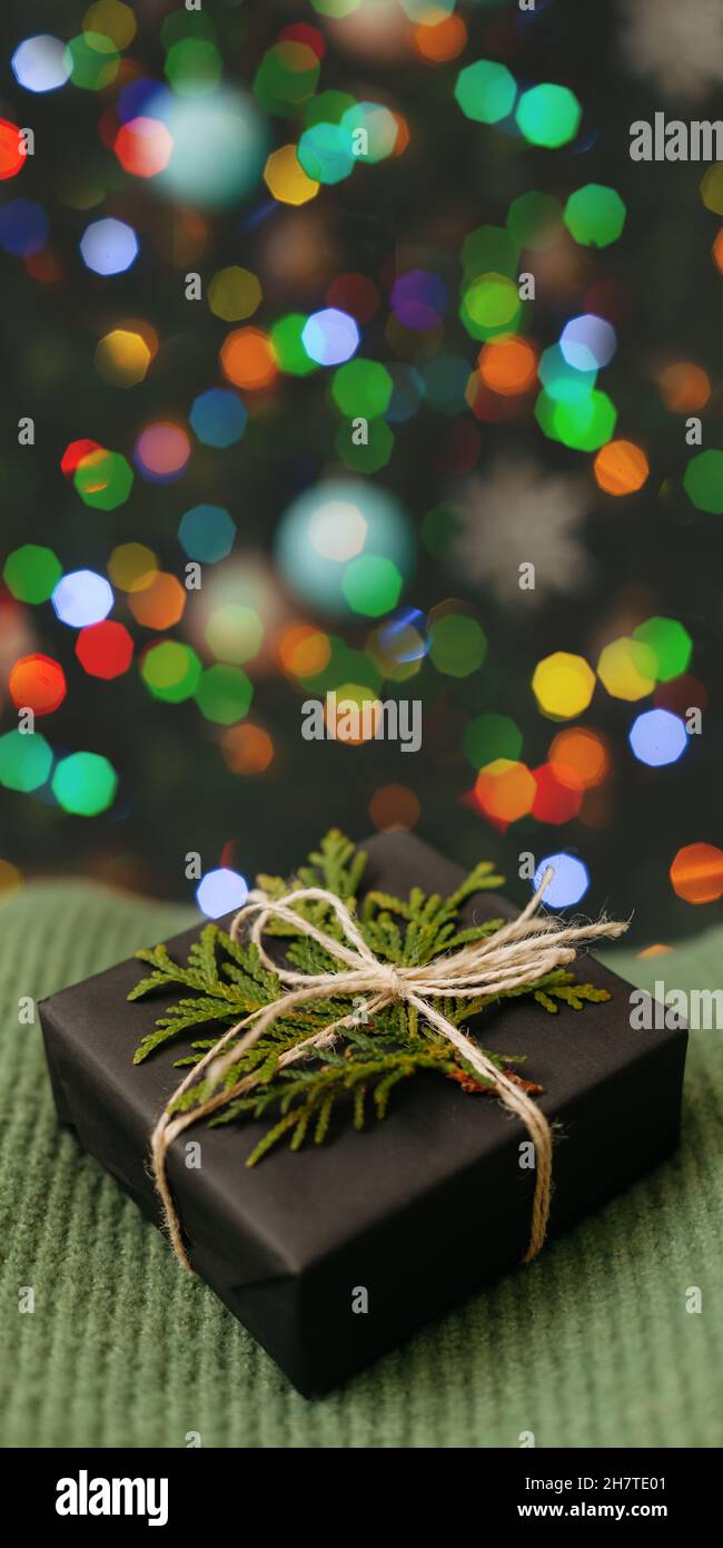 Schönes gemütliches Geschenk gewickeltes schwarzes Papier, das auf grünem Textil liegt, geschmückter Weihnachtsbaum auf Hintergrund. Helle Weihnachtskarte mit Platz für Text. Nahaufnahme Stockfoto