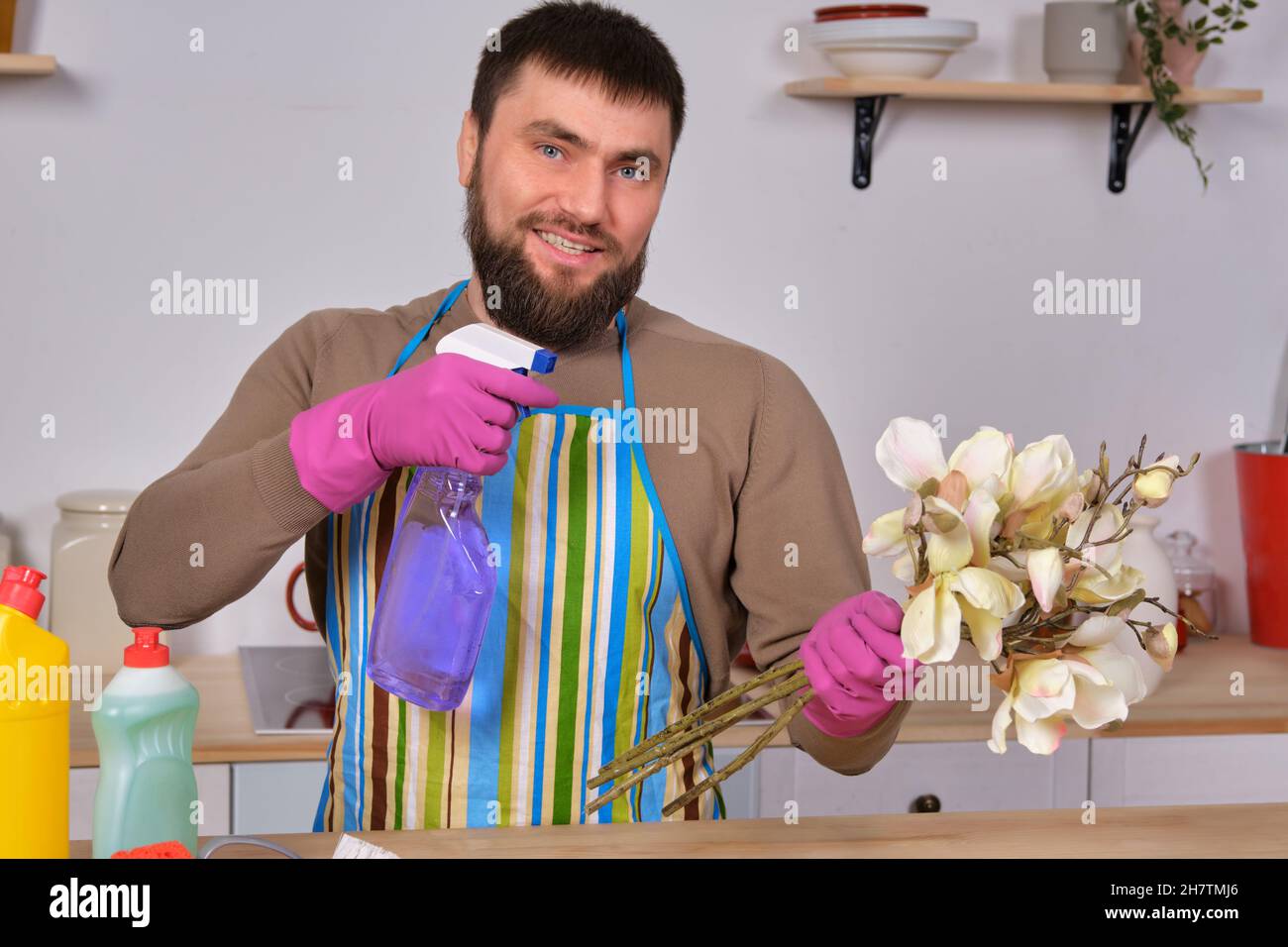 Junger, hübscher bärtiger Mann in der Küche zeigt alle seine Reinigungskräfte - Waschmittel, Bürsten, Sprays. Er denkt, er sei bereit für eine echte Reinigung Stockfoto