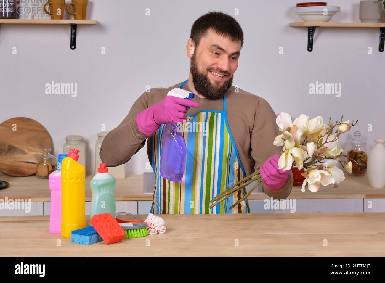 Junger, hübscher bärtiger Mann in der Küche zeigt alle seine Reinigungskräfte - Waschmittel, Bürsten, Sprays. Er denkt, er sei bereit für eine echte Reinigung Stockfoto