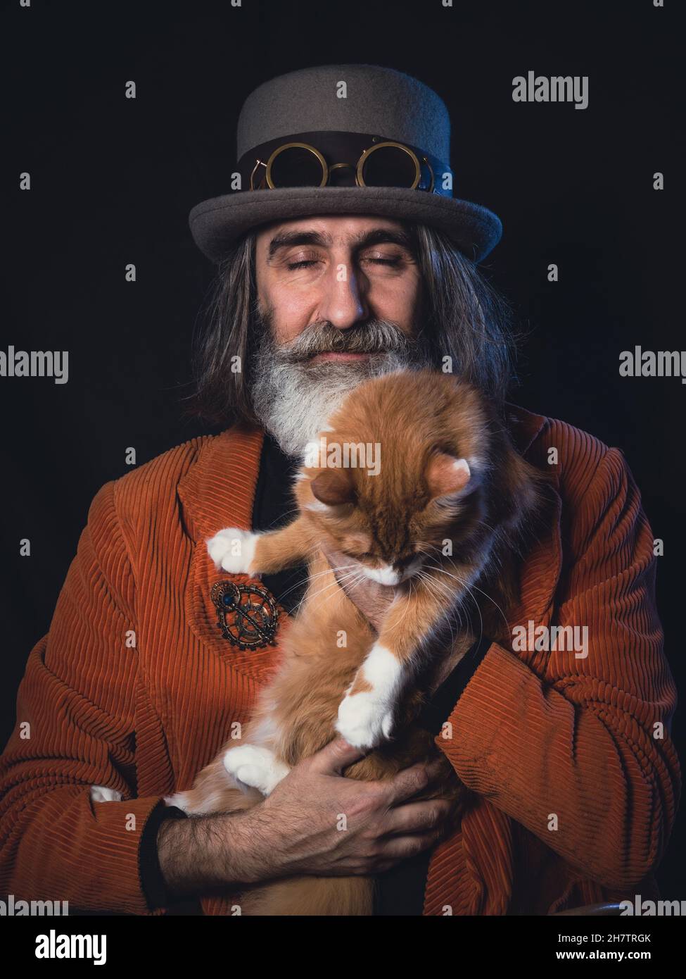 Ein grauhaariger bärtiger Intellektueller im reifen Alter in einem Steampunk-Hut hält eine Ingwerkatze, ein Mann ist glücklich, seine Augen sind geschlossen Stockfoto