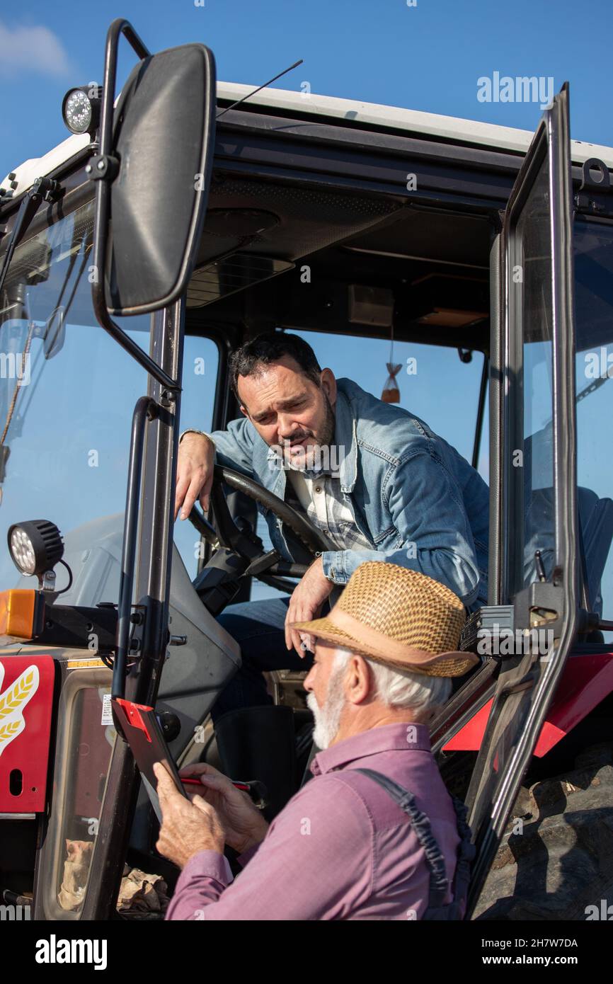 Reifer Fahrer, der im Traktor sitzt und neben dem Fahrzeug mit einem älteren Landwirt auf dem Feld spricht Stockfoto