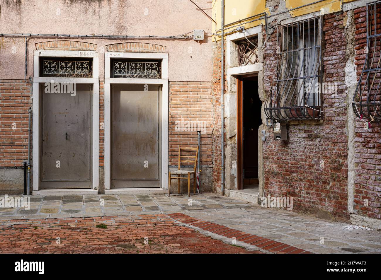 Blick auf eine kleine Gasse im Zentrum von Venedig. Ein Holzstuhl steht neben einer offenen Haustür. Stockfoto