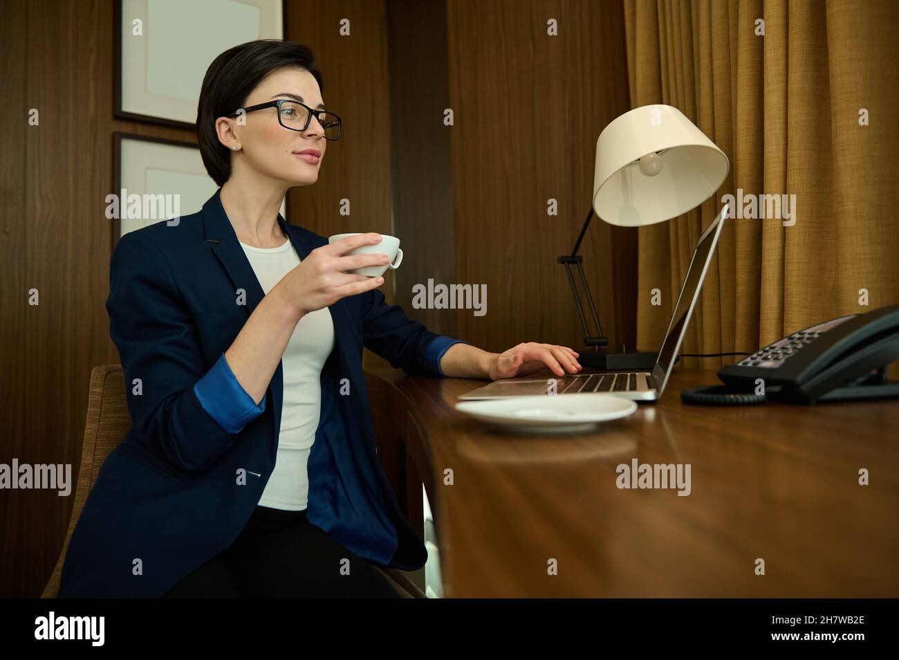 Junge Geschäftsfrau, Unternehmerin, erfolgreiche Vertriebsleiterin in Brillen und Business-Freizeitanzug sitzt am Schreibtisch und hält während der Arbeit eine Tasse Kaffee Stockfoto