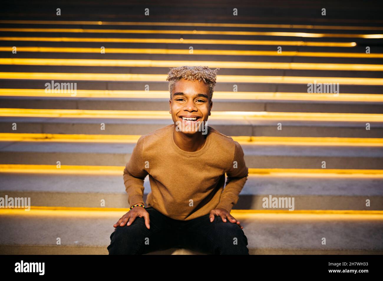 Junger lateinamerikanischer Mann sitzt auf der Treppe und lächelt auf der Straße vor der Kamera Stockfoto