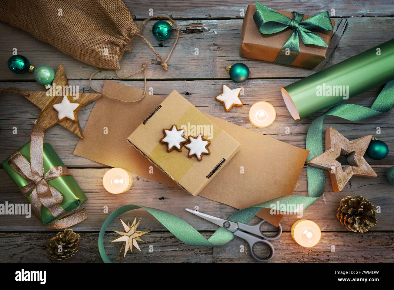 Feiertagsvorbereitung, Geschenkverpackung mit Naturpapier und grünem Band, Weihnachtsdekoration wie Kugeln, Sterne und brennende Kerzen auf einer rustikalen Holzlasche Stockfoto