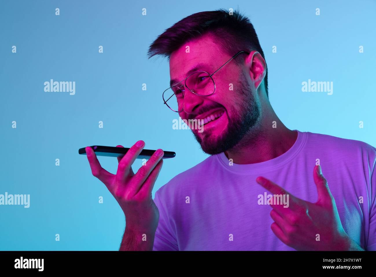 Beschnittenes Porträt eines aufgeregten Mannes, der fröhlich Sprachnachrichten aufzeichnete, wobei das Telefon auf blauem Hintergrund in Neonlicht isoliert war Stockfoto