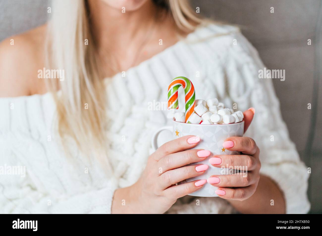 Blonde Frau trägt ein weißes Strickkleid und hält einen Becher mit heißem Kakao mit Marschmalchen und Zuckerrohr. Weihnachtsstimmung Stockfoto