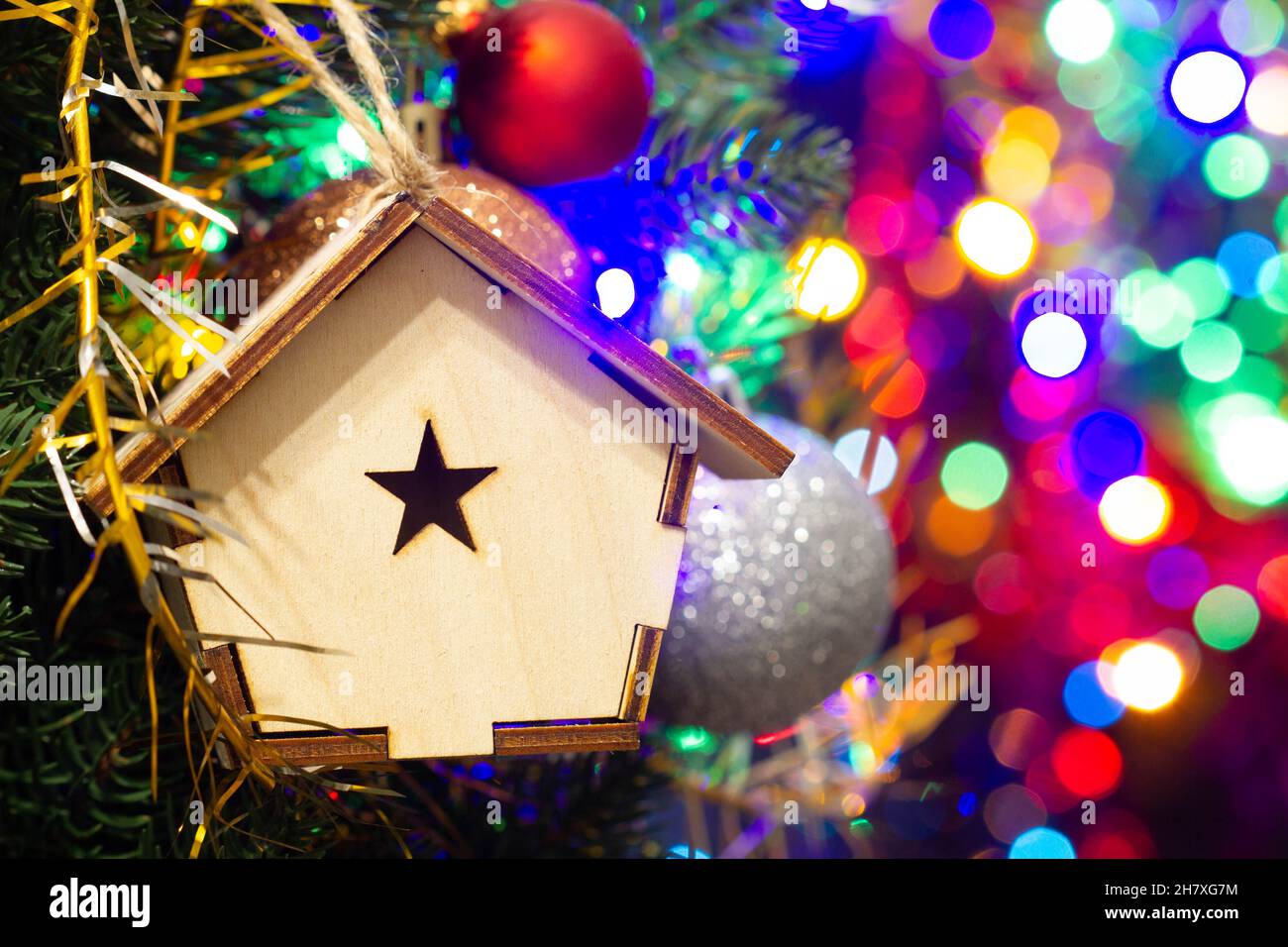 Weihnachtsspielzeug Holzhaus auf dem Weihnachtsbaum auf der bunten Beleuchtung Hintergrund. Wunderschöne Weihnachtsdekorationen. Coole helle Neujahrsspielzeuge Stockfoto