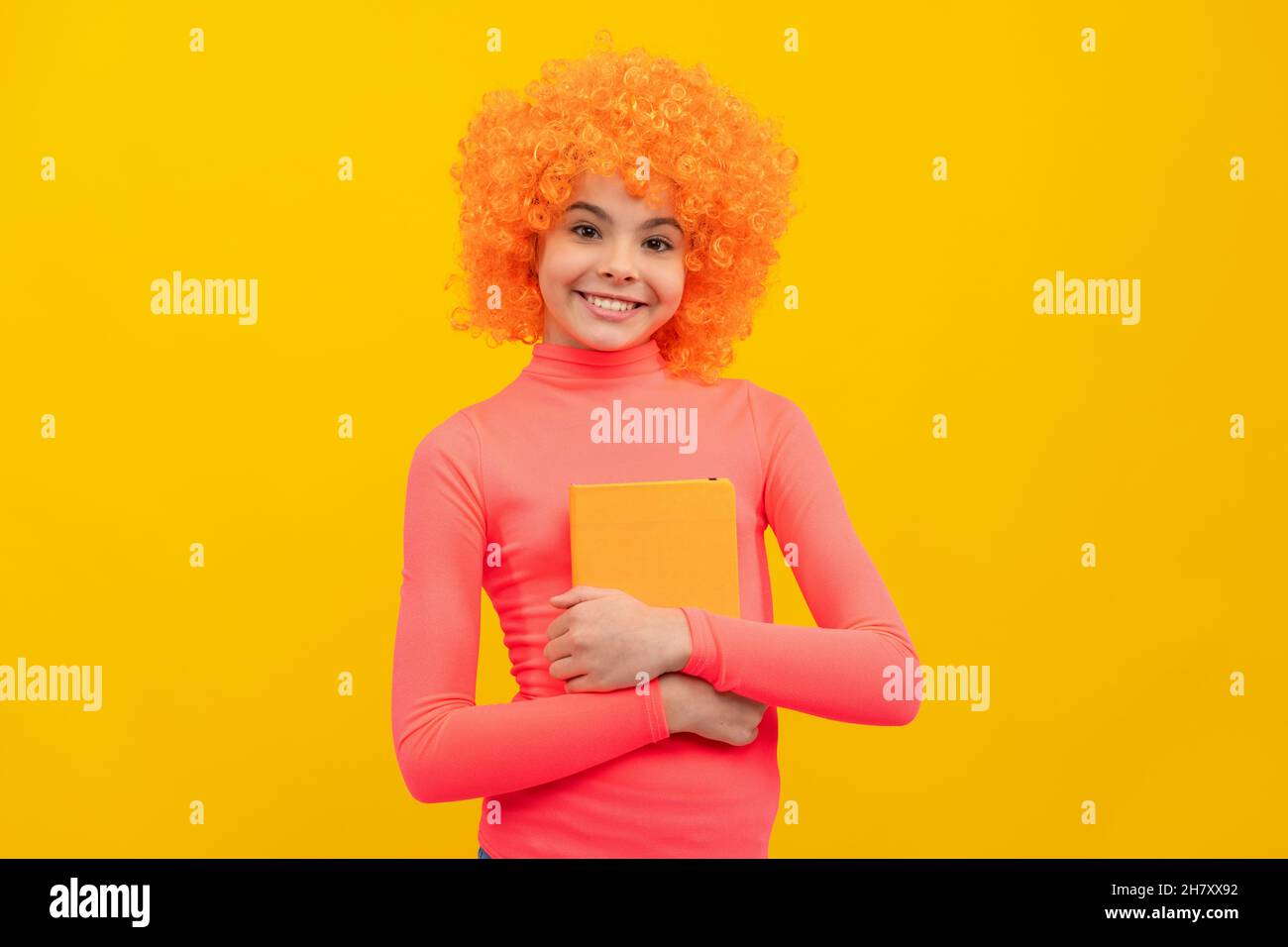 Glückliches Mädchen Kind mit orangen Haaren in rosa Poloneck Lächeln halten Buch gelben Hintergrund, Schule Stockfoto