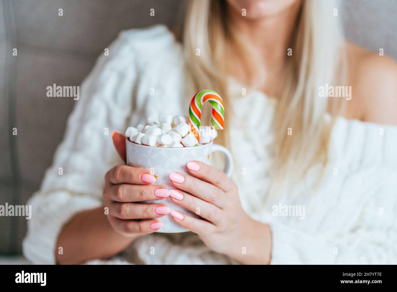 Blonde Frau trägt ein weißes Strickkleid und hält einen Becher mit heißem Kakao mit Marschmalchen und Zuckerrohr. Weihnachtsstimmung Stockfoto
