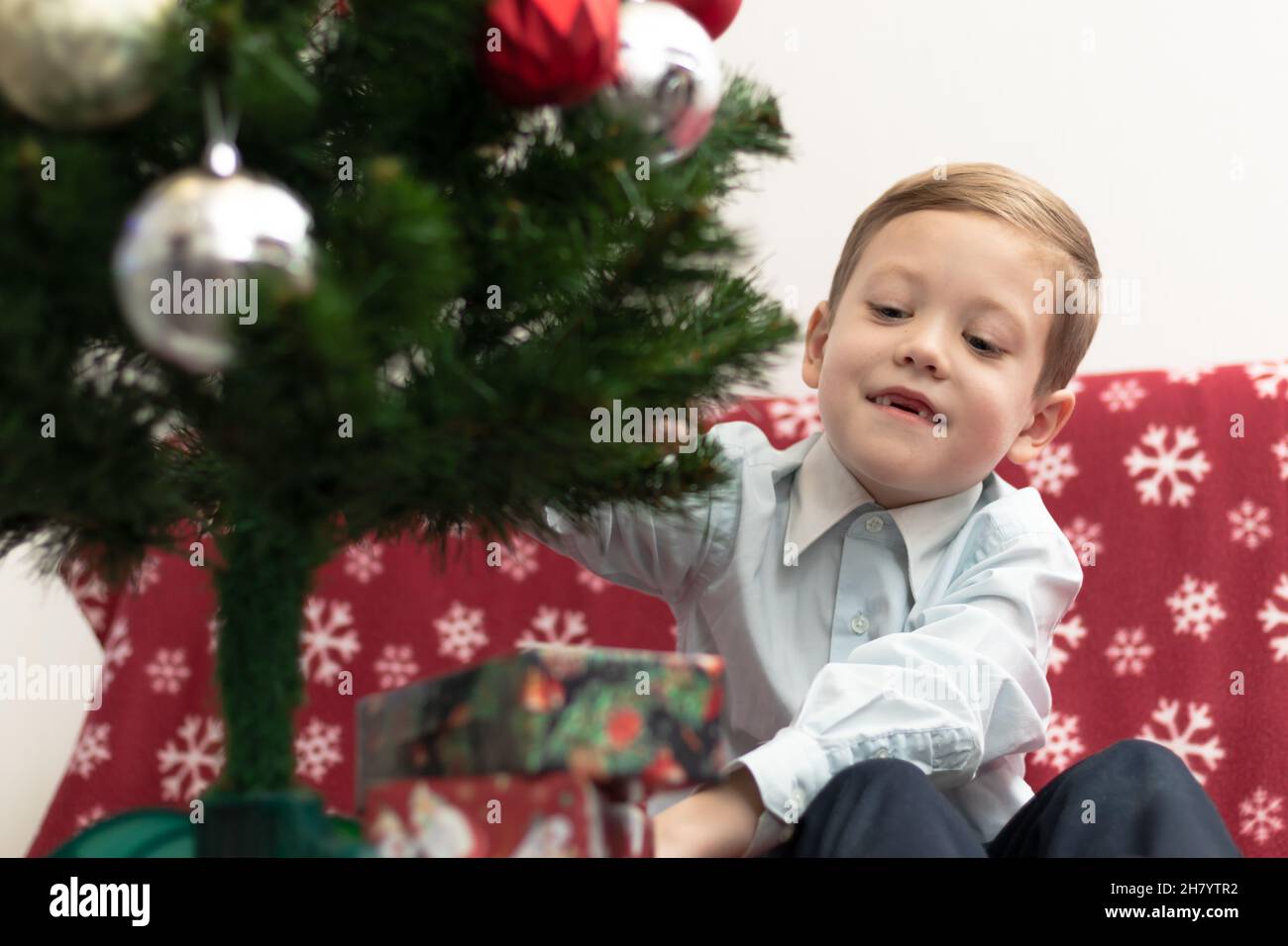 Ein siebenjähriger Junge freut sich vor dem Hintergrund eines Neujahrsbaums, der auf einer warmen Decke sitzt, über Geschenke für das neue Jahr. Selektiver Fokus. Portra Stockfoto