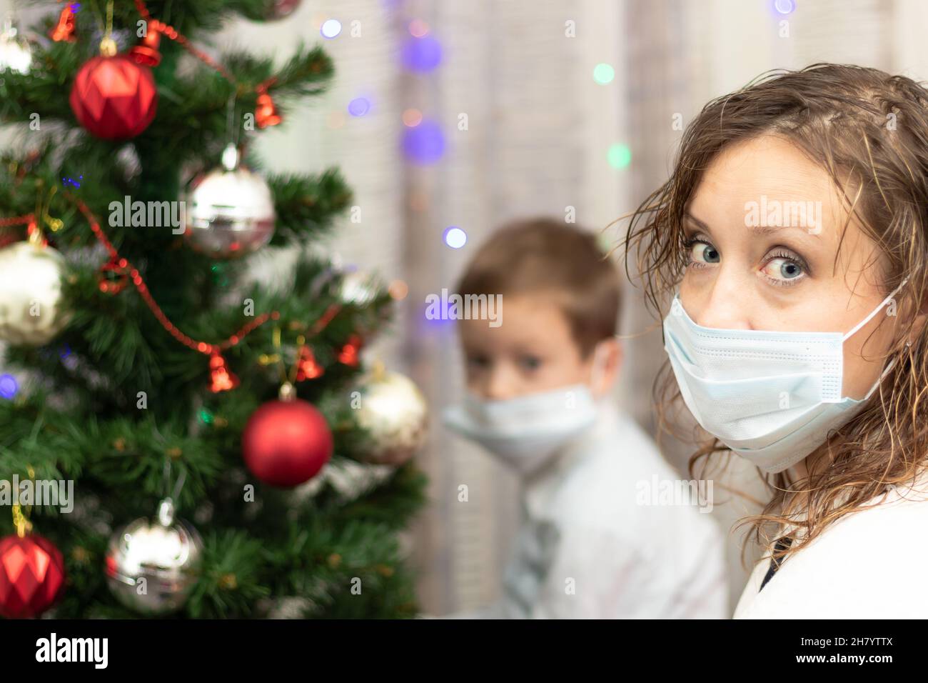 Mutter und Sohn in medizinischen Masken am Weihnachtsbaum zu Hause während der Coronavirus-Pandemie am Silvesterabend. Selektiver Fokus. Hochformat Stockfoto