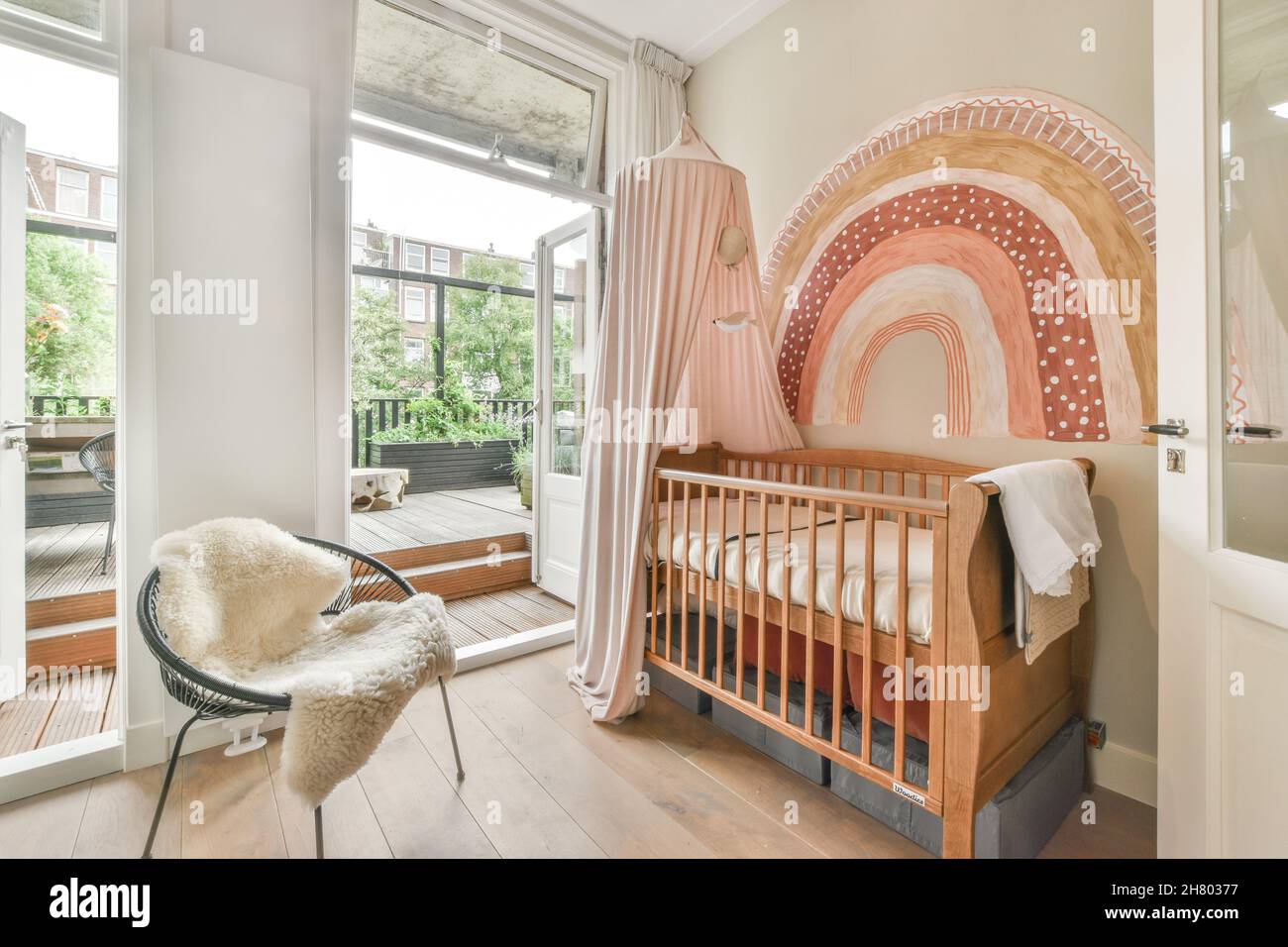 Holzkrippe in der Nähe der Wand mit dekorativem Element in der Nähe von Sessel mit karierten in hellen Schlafzimmer mit Terrasse mit Blick auf die Stadt mit Bäumen platziert Stockfoto