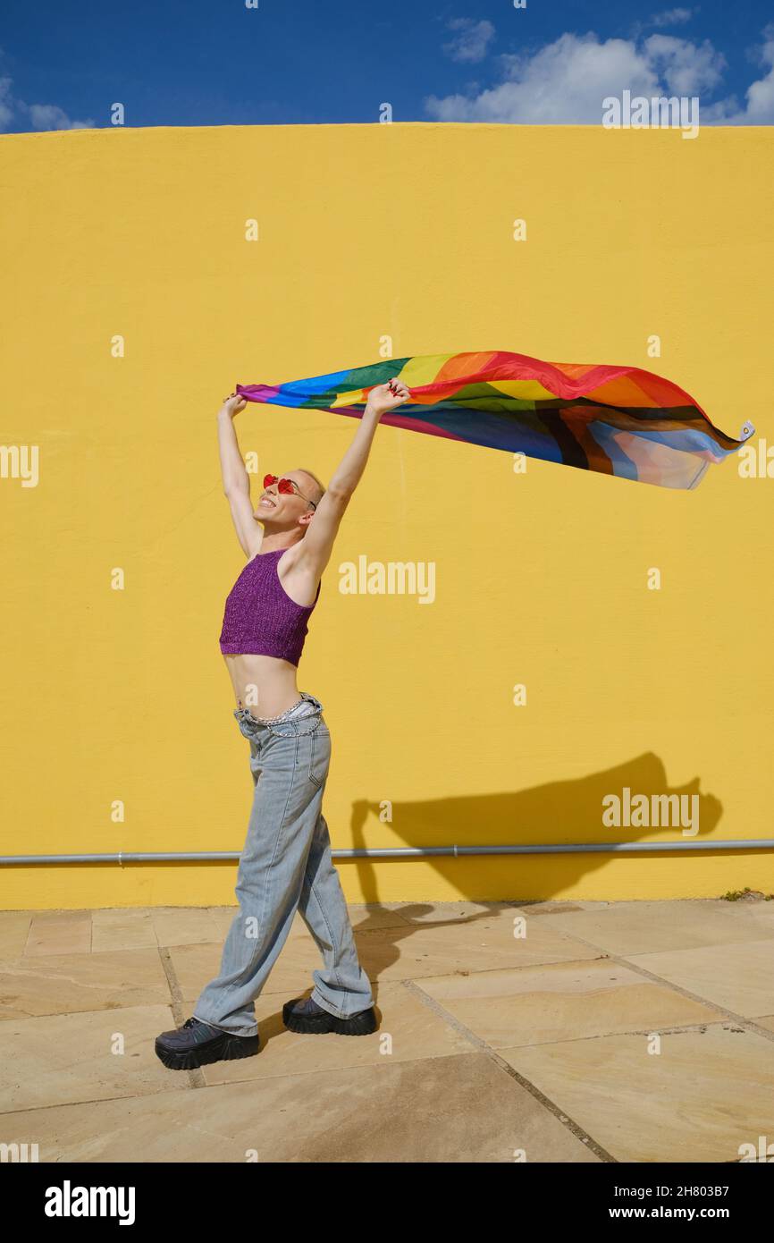Glückliche junge nicht-binäre Person hält und winkt eine Regenbogenfahne im Freien. Stockfoto