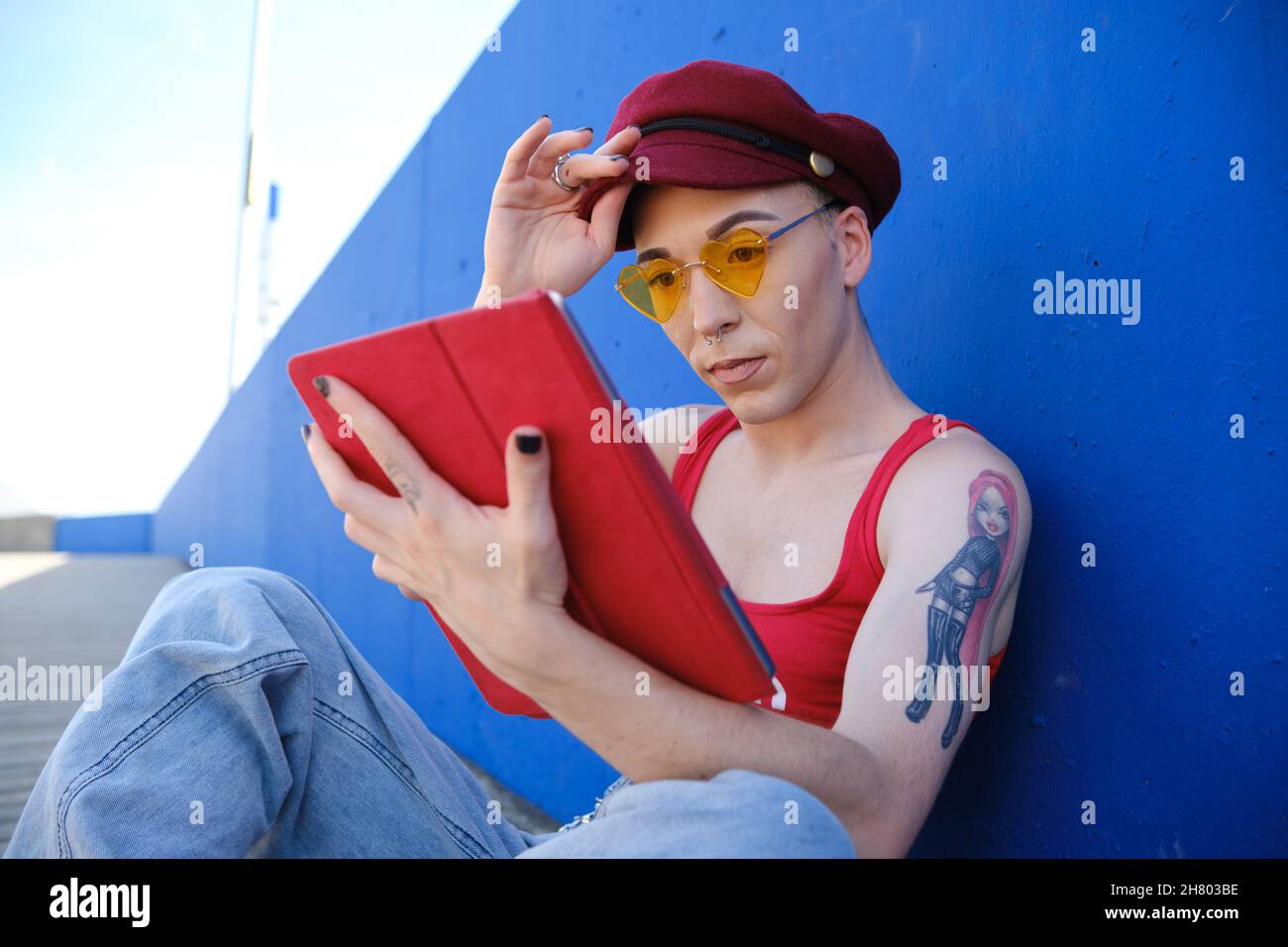 Junge nicht-binäre Person, die ein digitales Tablet benutzt, während sie entspannt auf der Straße sitzt. Stockfoto