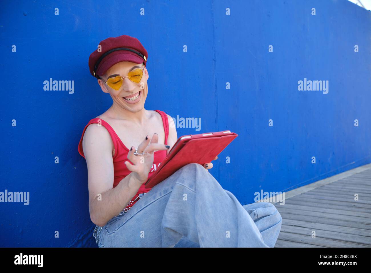 Lächelnder junger, nicht-binärer Mann in Baskenmütze und Sonnenbrille mit einem digitalen Tablet, während er im Freien sitzt. Stockfoto
