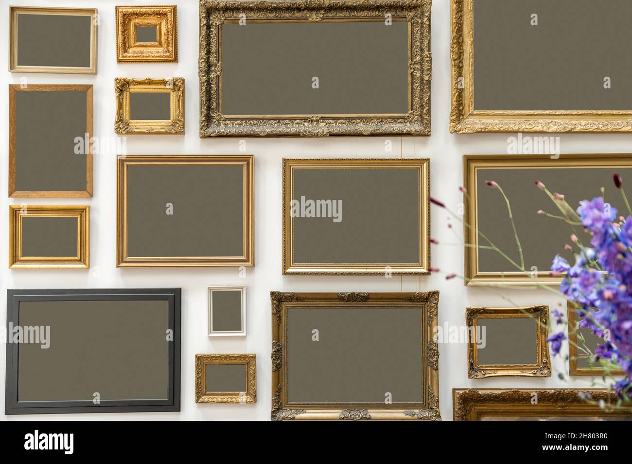 Sammlung von verschiedenen leeren Bilderrahmen mit goldenen Rändern in verschiedenen Größen, die an der weißen Wand im Raum mit Blumen hängen Stockfoto