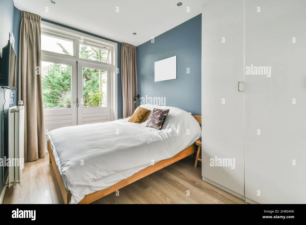 Komfortables Bett mit weißer Decke und Kissen neben dem Kleiderschrank und dem modernen Fernseher im hellen Schlafzimmer mit Fenster im Apartment Stockfotografie