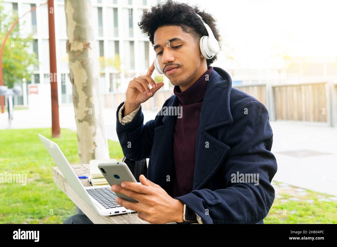 Konzentrierter afroamerikanischer Student im Mantel, der Musik in kabellosen Kopfhörern hört, während er im Park auf dem Handy surft und auf dem Laptop studiert Stockfoto