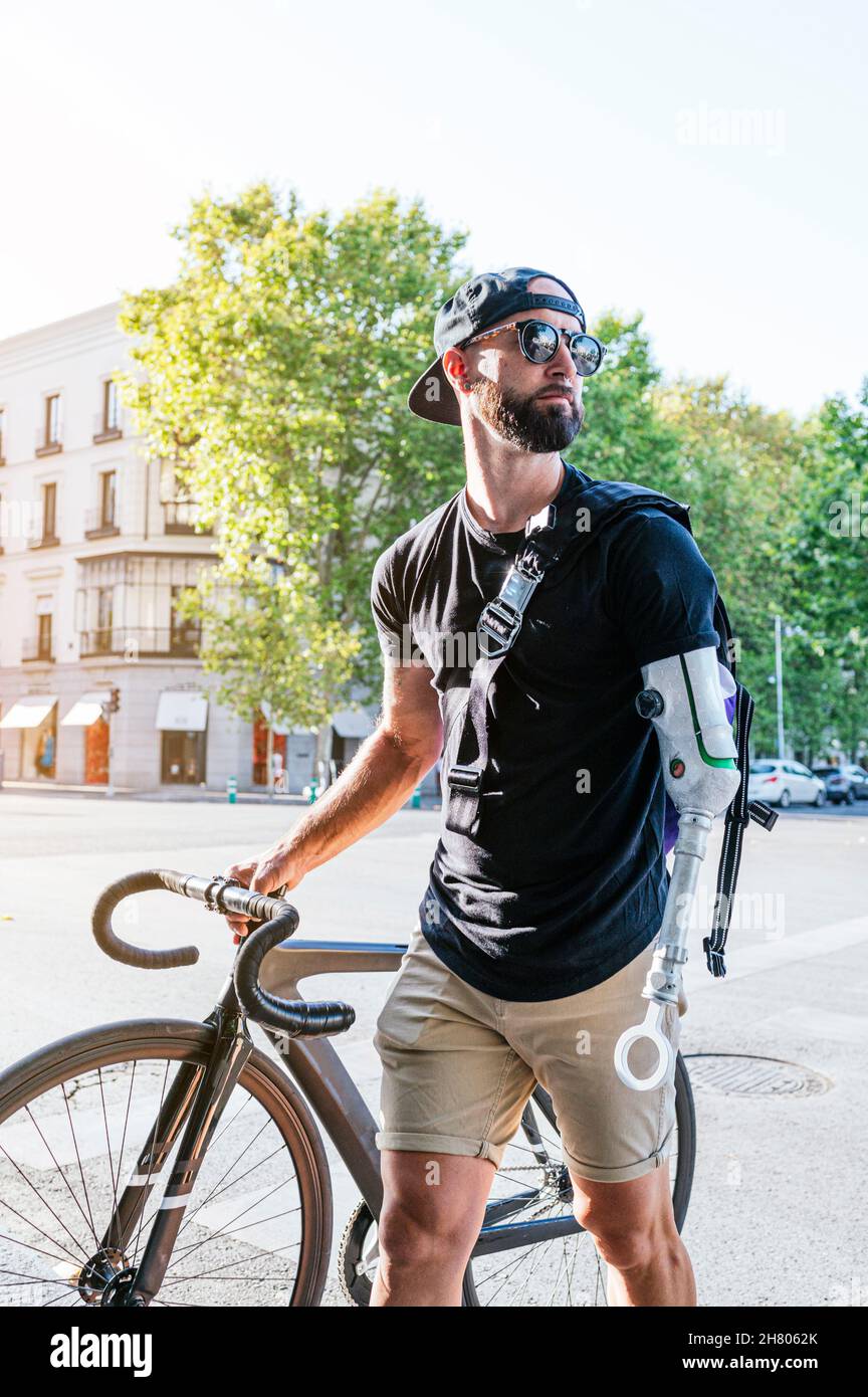Männlicher Hipster mit Armprothese, der mit dem Fahrrad auf dem Bürgersteig in der Stadt läuft Stockfoto