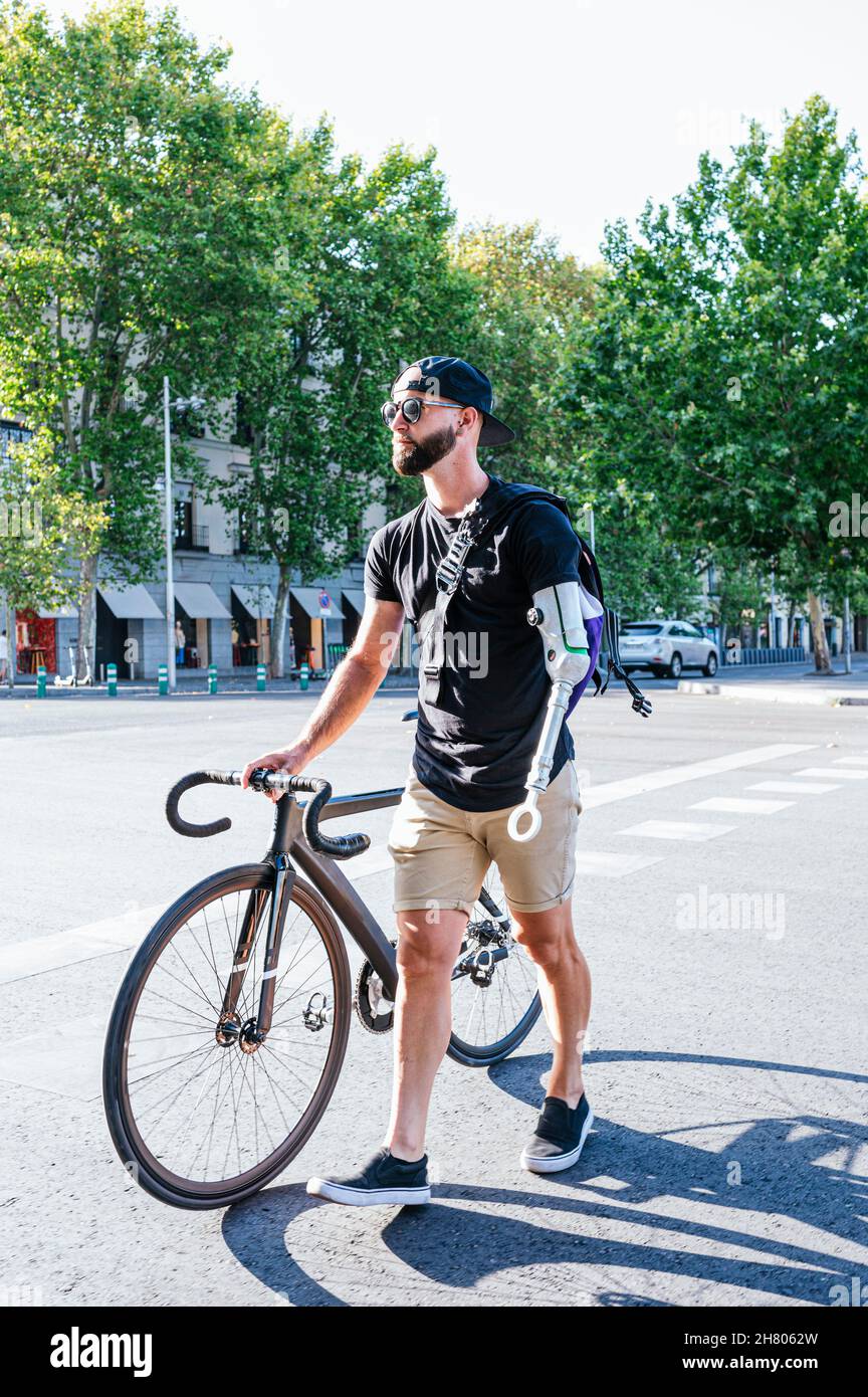 Männlicher Hipster mit Armprothese, der mit dem Fahrrad auf dem Bürgersteig in der Stadt läuft Stockfoto