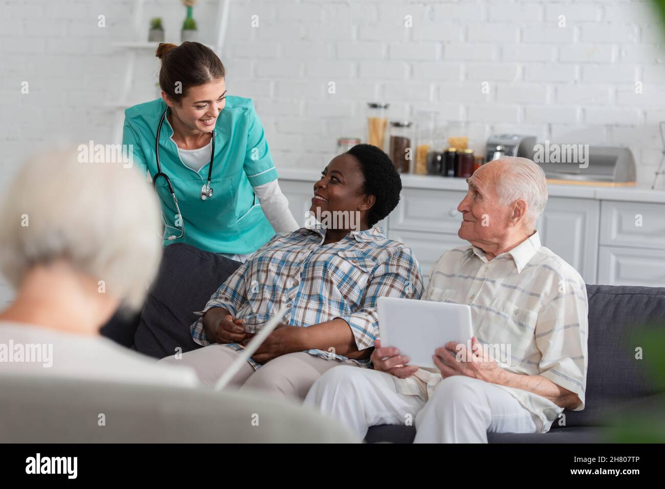 Positive Krankenschwester, die mit afroamerikanischer Frau mit Tee in der Nähe von älteren Patienten im Pflegeheim spricht Stockfoto