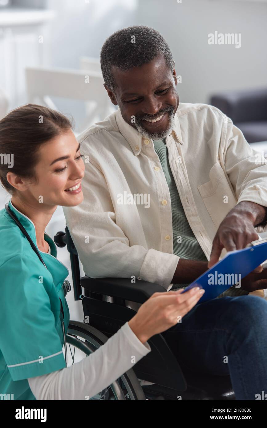 Lächelnde junge Krankenschwester hält Zwischenablage in der Nähe von afroamerikanischen Patienten im Rollstuhl im Pflegeheim Stockfoto