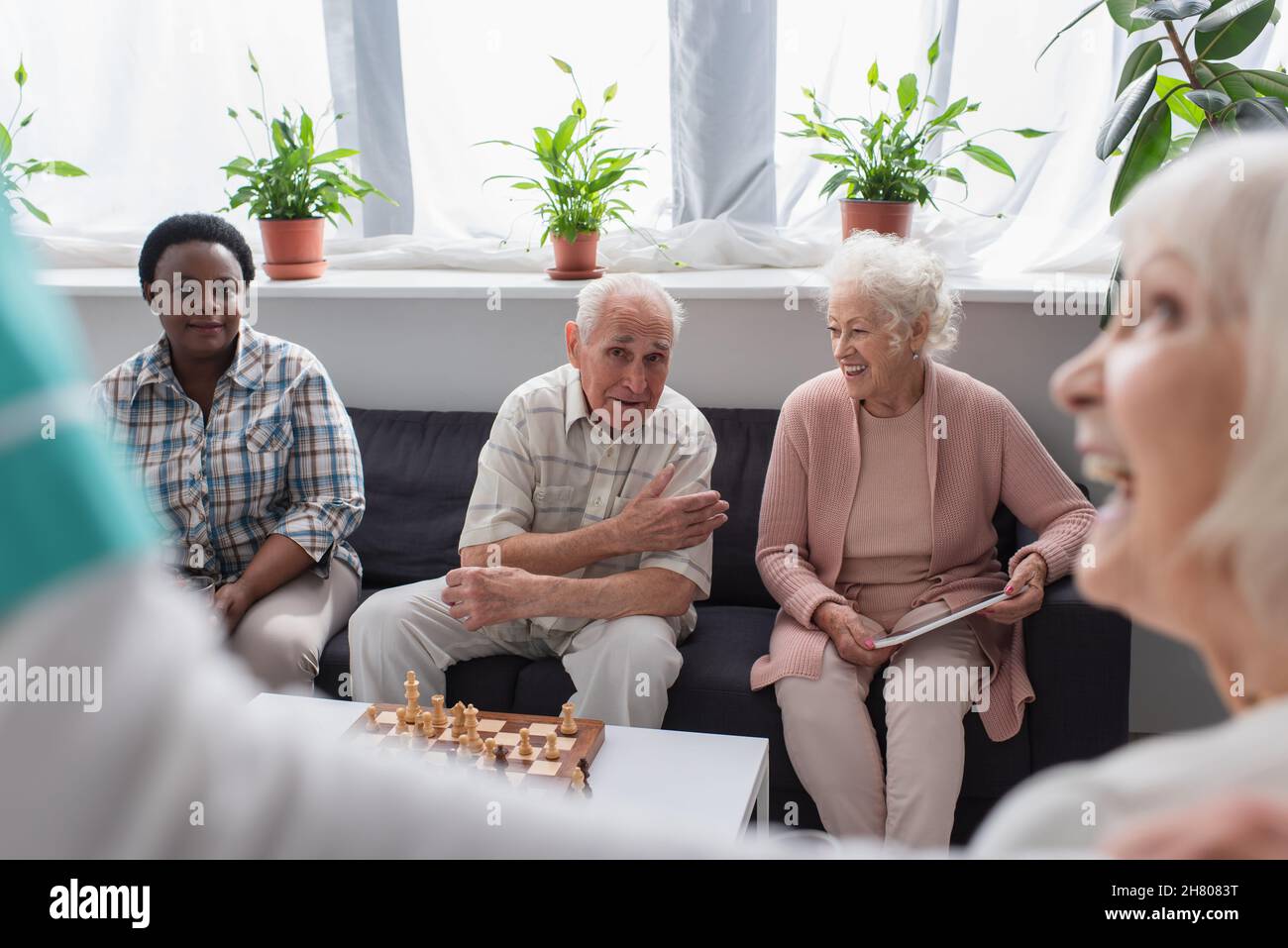 Ältere interrassische Menschen mit digitalem Tablet, die in der Nähe von Schach sprechen, und verschwommene Krankenschwester im Pflegeheim Stockfoto