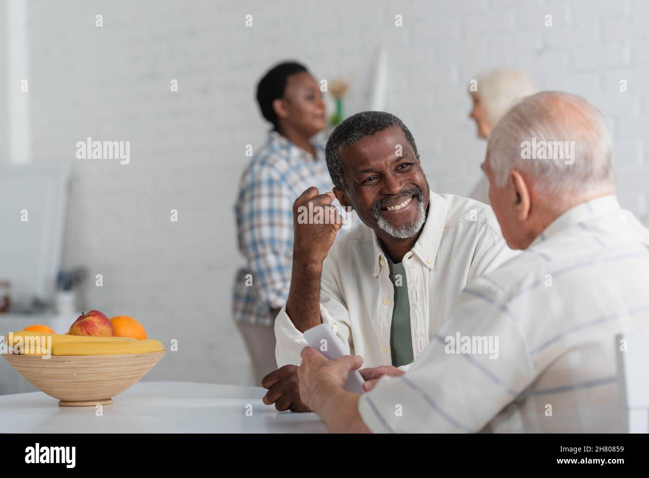 Lächelnder afroamerikanischer Mann mit Ja-Geste in der Nähe eines älteren Freundes mit Smartphone und verschwommenen Frauen im Pflegeheim Stockfoto
