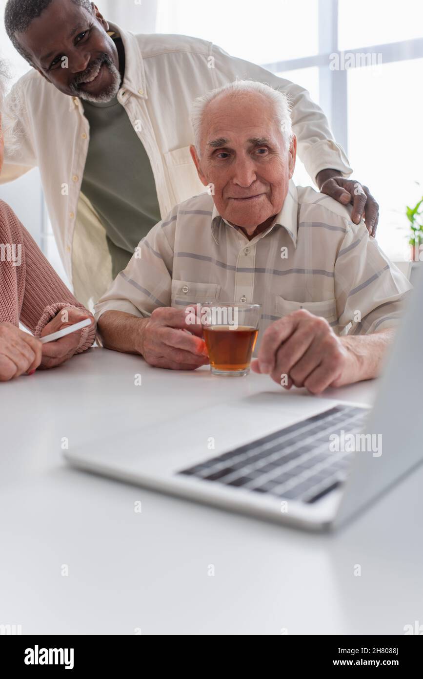 Lächelnder afroamerikanischer Mann umarmt ältere Freunde mit Tee in der Nähe eines Laptops im Pflegeheim Stockfoto
