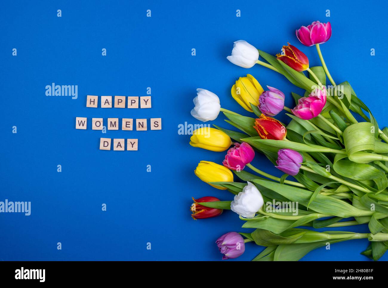 Happy Women Tageskarte. Holzbuchstaben. Mix aus Frühlingsblumen. Blauer Hintergrund mit Blumen Tulpen close-up verschiedenen Farben. Mehrfarbige Frühlingsblume. Geschenk. Rot, Rosa, Weiß und Gelb. Stockfoto