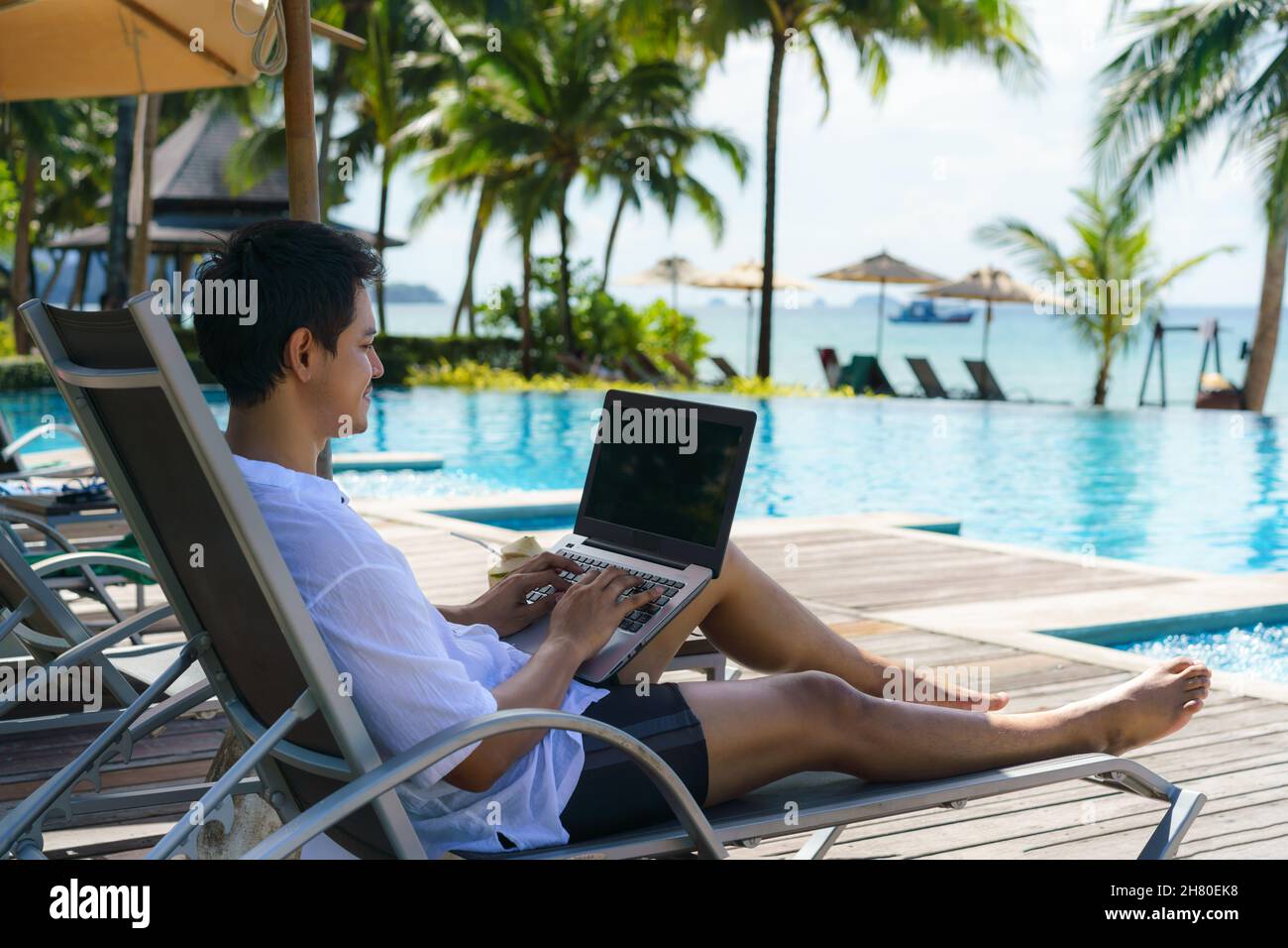 Der asiatische Mann verbrachte seinen Sommerurlaub mit der Arbeit an seinem Laptop in einem Stuhl in der Nähe des Swimmingpools im Resorthotel in der Nähe des Meeres. Stockfoto Der asiatische Mann verbrachte seinen Sommerurlaub mit der Arbeit an seinem Laptop in einem Stuhl in der Nähe des Swimmingpools im Resorthotel in der Nähe des Meeres. Stockfoto