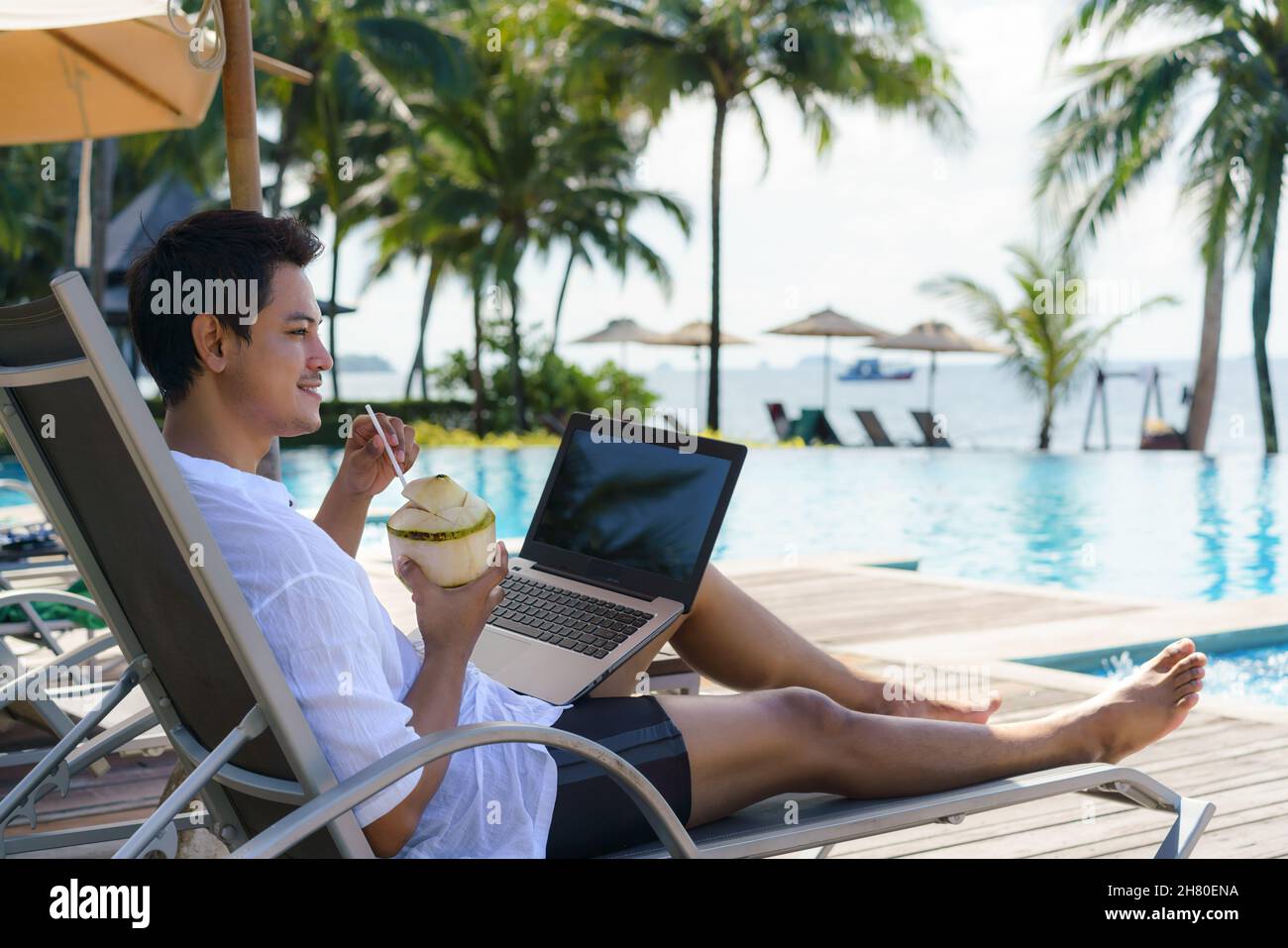 Der Asiate trinkt Kokoswasser, während er in den Sommerferien an seinem Laptop auf einem Sessel am Pool im Resorthotel arbeitet. Stockfoto Der Asiate trinkt Kokoswasser, während er in den Sommerferien an seinem Laptop auf einem Sessel am Pool im Resorthotel arbeitet. Stockfoto