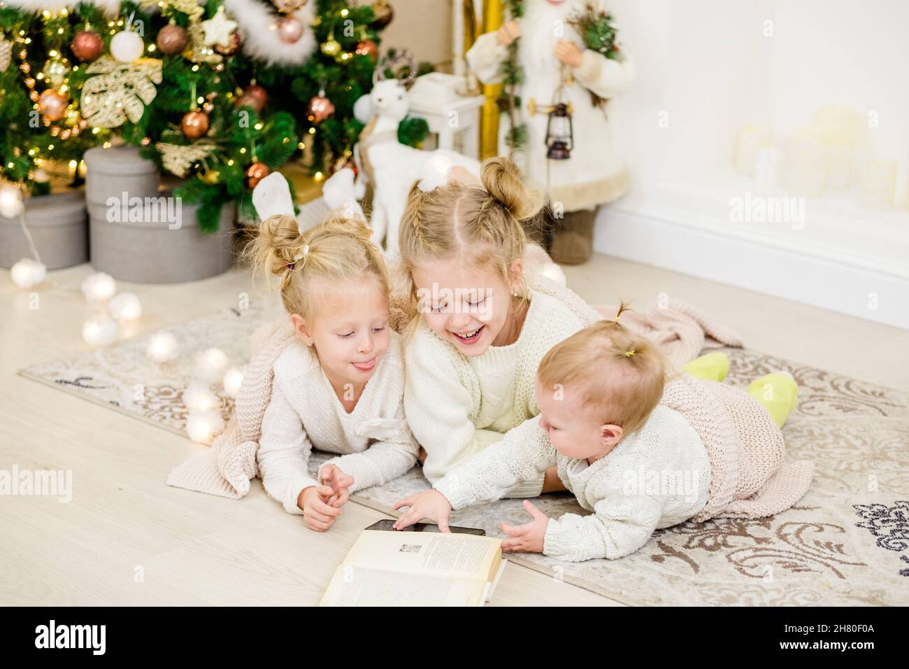 3 blonde Mädchen liegen am Weihnachtsbaum auf dem Boden und lesen ein Buch. Gemütliche Abende mit einem Buch am Baum. Freundinnen lesen zusammen ein Buch. Stockfoto