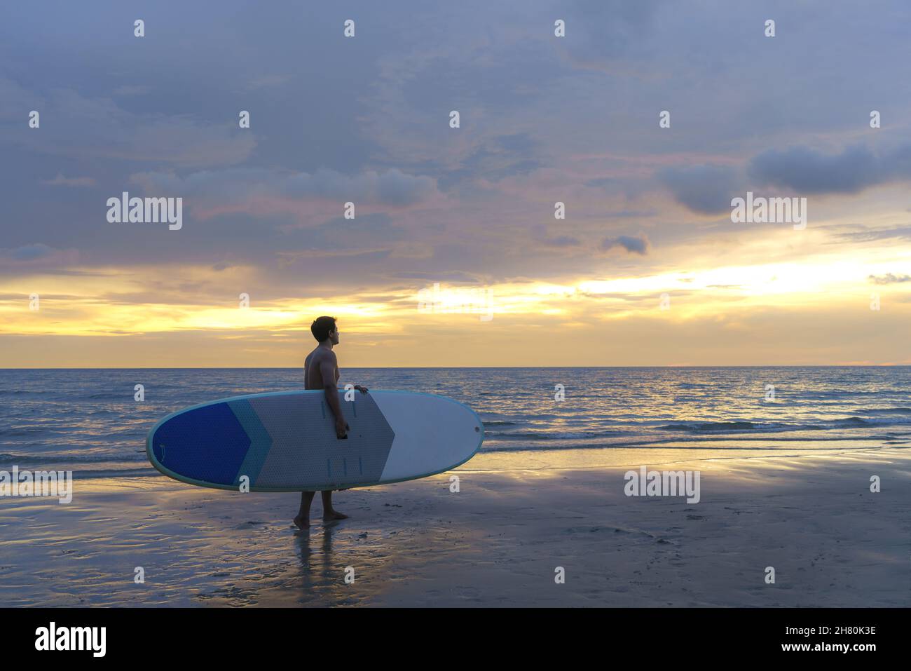 Asiatischer Mann hält ein Brett und paddelt und läuft am Strand. Im Hintergrund das Meer und der Sonnenuntergang. Rückansicht. Surfen im Sommer. Stockfoto