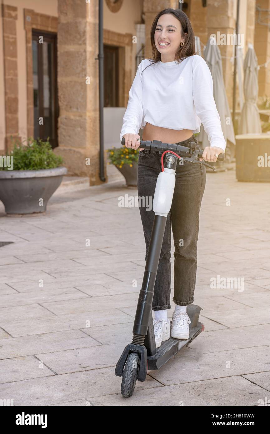 Junge glückliche braune Frau, die in der Stadt auf einem Elektroroller reitet und die Kamera anlächelt Stockfoto