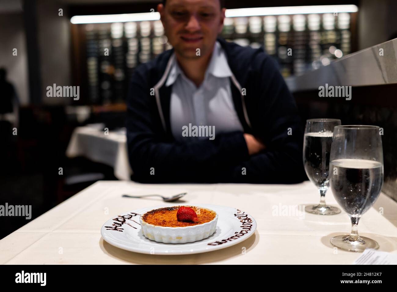 Junger glücklicher Mann, der zum Abendessen im Restaurant saß und das Essen auf dem Date mit französischem Creme-Brulee-Dessert und einem Schild zum Happy Anniversary ansah Stockfoto