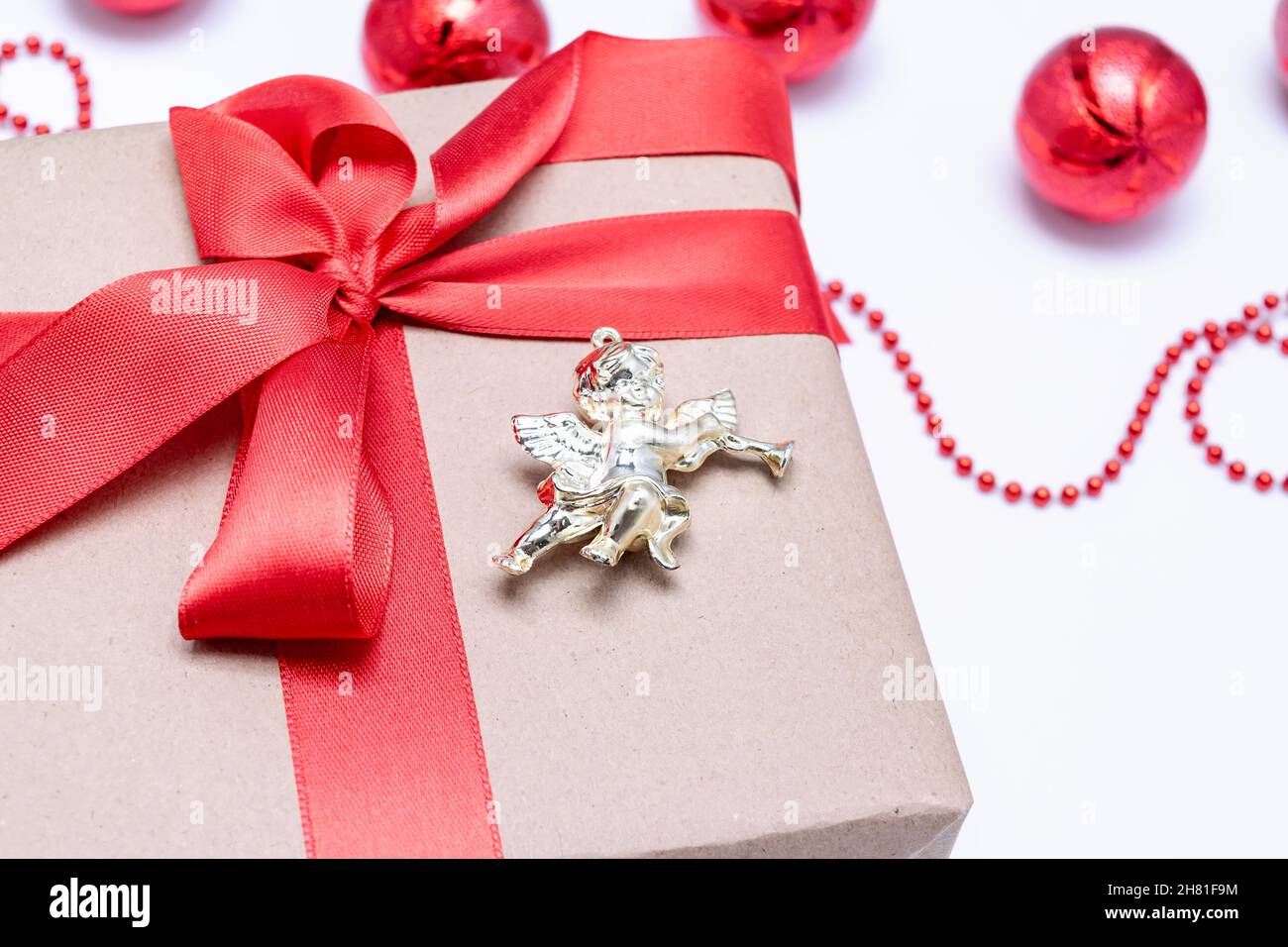 Nahaufnahme eines Weihnachtsgeschenks mit einem roten Band und einem Engel neben den roten Weihnachtskugeln auf weißem Hintergrund. Festliche Postkarte. Horizontales Foto. Stockfoto