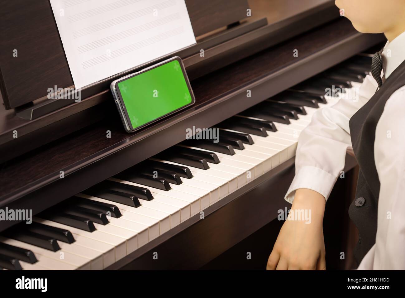 Ein Junge sitzt an einem elektronischen Klavier und schaut in ein Smartphone mit grünem Bildschirm. Modell für mobile Musik-Apps. Klavierunterricht online. Online-Musikerziehung Stockfoto