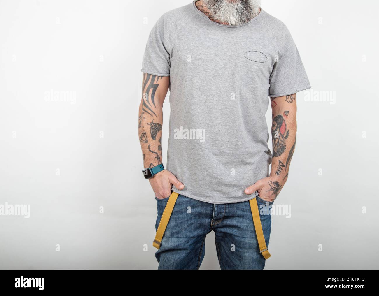 Bärtig tätowierte Mann posiert in Jeans mit Hosenträgern und grauem Kurzarm-T-Shirt aus Baumwolle, auf weißem Hintergrund. Stockfoto