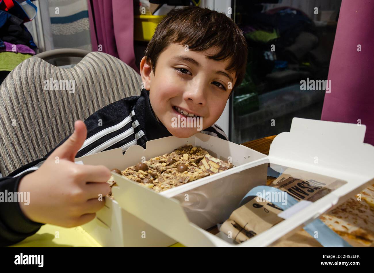 Ein Junge gibt seinen Daumen nach oben Zustimmung und lächelt auf die Kamera als Reaktion auf das Öffnen einer Kiste mit Tiffin. Stockfoto