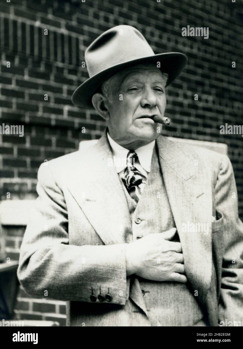 Alter Mann in einer städtischen Umgebung mit Zigarre, Fedora-Hut und Anzug, um 1950. Stockfoto