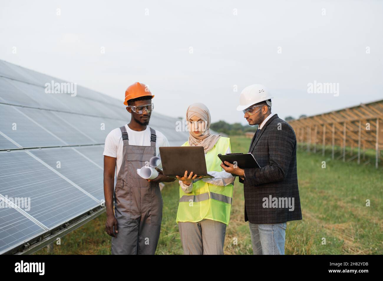 Muslimische Frau in Hijab und indischer Mann in weißem Helm im Gespräch mit afroamerikanischem Techniker auf Solarstation. Menschen mit mehreren Rassen, die Laptops, Clipboard und Blaupausen für die Arbeit im Freien verwenden. Stockfoto